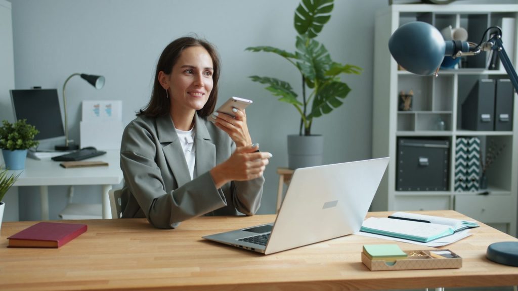 Person in a blazer using a smartphone beside an open laptop at an office desk.